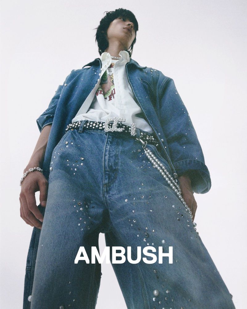 Stylish person poses in denim jacket and jeans with pearl accents against a bright background, showcasing high-fashion street style.