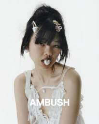 Close-up portrait of an East Asian woman with a white heart-shaped accessory in her mouth, wearing white top and pearl accessories against a white background.