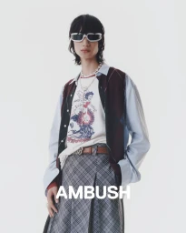 Model wearing graphic t-shirt, argyle vest, light blue shirt, plaid skirt, and white sunglasses against a white background.