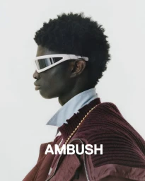 Profile portrait of a Black man wearing white wrap-around sunglasses and a maroon layered knit collar against a plain background