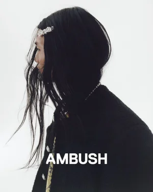 Profile portrait of a model with long dark hair wearing a jeweled hair clip and black jacket against a white background for Ambush SS25.