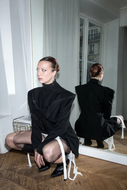 Woman in black jacket crouching indoors, reflected in mirror, herringbone floor, fair skin, looking left, high fashion.