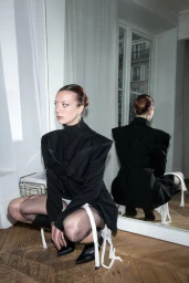 Woman in a structured black coat and sheer tights squatting in a bright room with a large mirror.