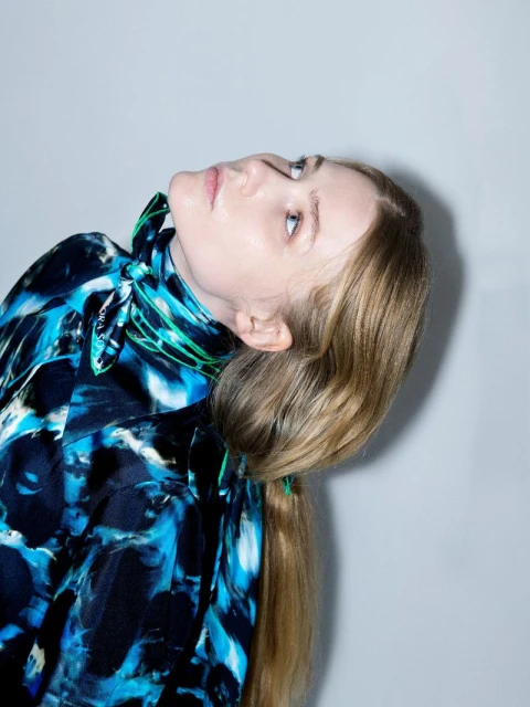 Woman with blonde hair and patterned shirt looks up in a fashion-style portrait on a neutral grey background with studio lighting.