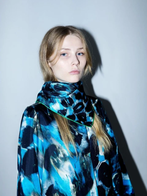 Portrait of a blonde woman wearing a blue patterned top, centered against a neutral background in a fashion-style photograph.