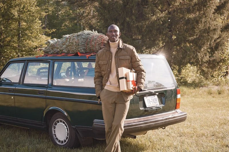 An African-American man carrying gift boxes stands next to a dark green station wagon with a Christmas tree strapped to its roof.