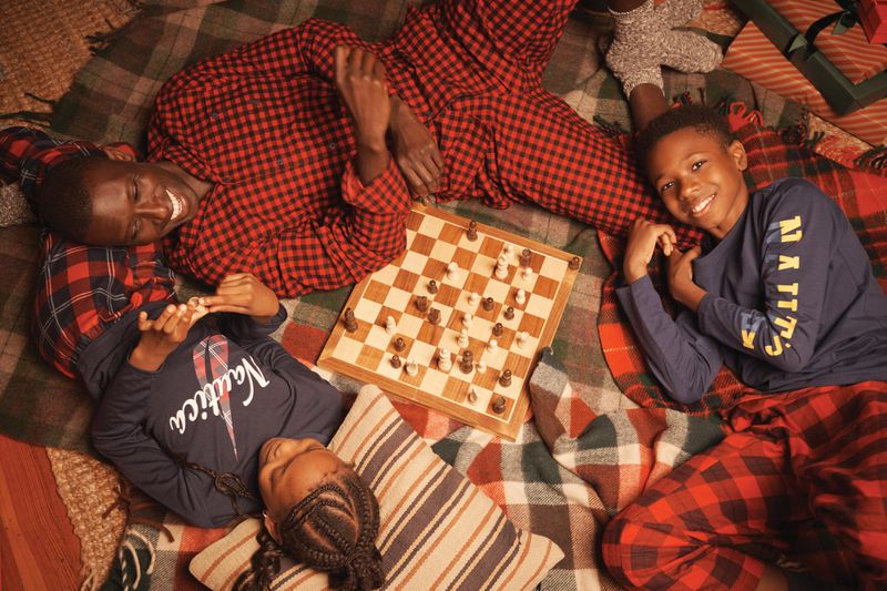 Overhead shot of three children and an adult playing chess on a blanket, all in plaid pajamas, indoors, with warm lighting.