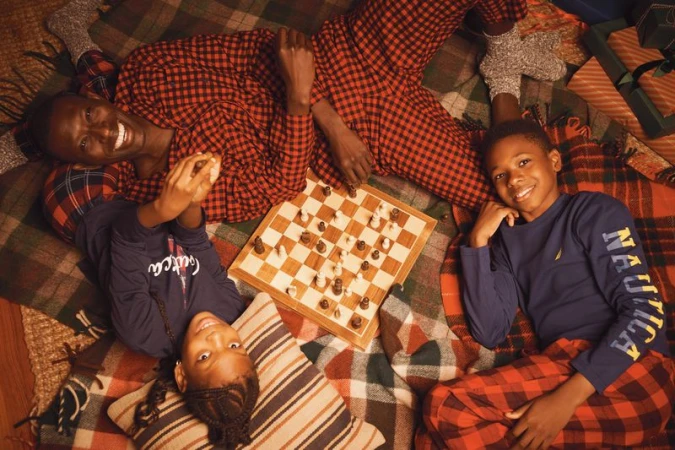 African-American family of three smiles while playing chess on a plaid blanket, surrounded by gifts and wearing matching pajamas.