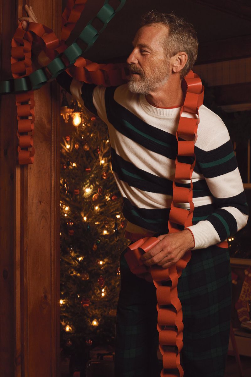 Adult male with grey hair decorates a Christmas tree with a handmade red and green paper chain in a cozy, warmly-lit indoor environment.