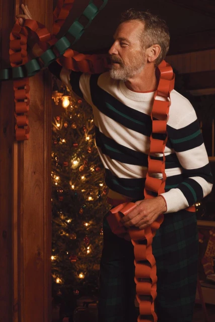 Adult male with grey hair decorates a Christmas tree with a handmade red and green paper chain in a cozy, warmly-lit indoor environment.
