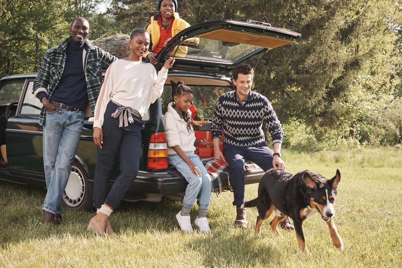 A family of five, including parents, children, and a dog, pose near a vintage car in a grassy, wooded area, wearing casual, seasonal attire.