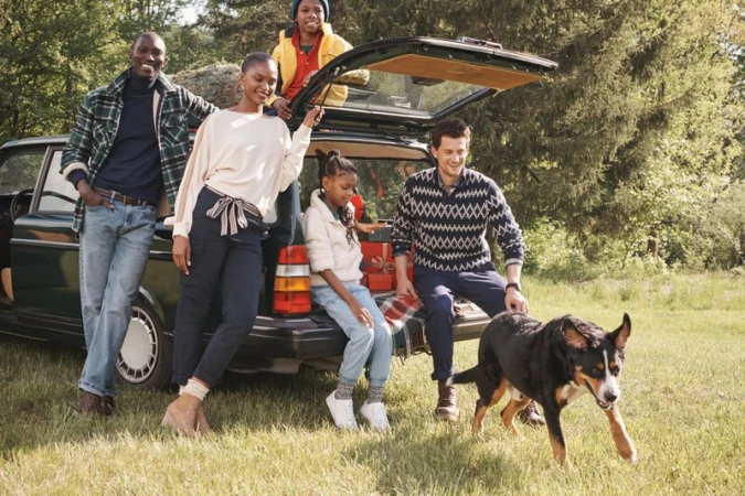 A family of five, including parents, children, and a dog, pose near a vintage car in a grassy, wooded area, wearing casual, seasonal attire.