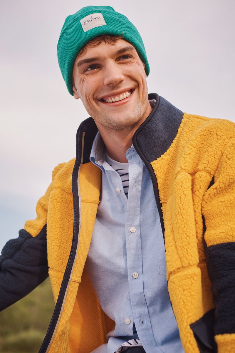 Smiling man in green beanie and yellow fleece jacket, outdoor apparel advertisement promoting fashion and lifestyle in natural lighting.
