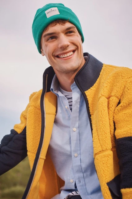 Smiling man in green beanie and yellow fleece jacket, outdoor apparel advertisement promoting fashion and lifestyle in natural lighting.