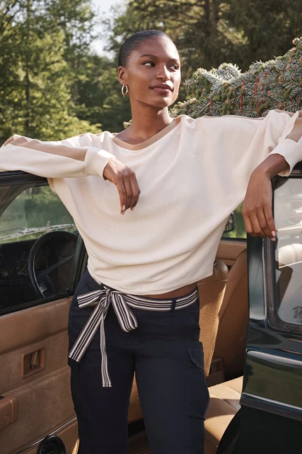 Stylish woman in cream sweater and navy pants poses next to a vintage car in a lush, green, natural outdoor setting on a bright day.