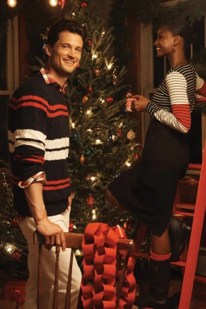 Smiling man stands near a Christmas tree while an adult female decorates with gift. Indoor holiday scene with red and green colors.