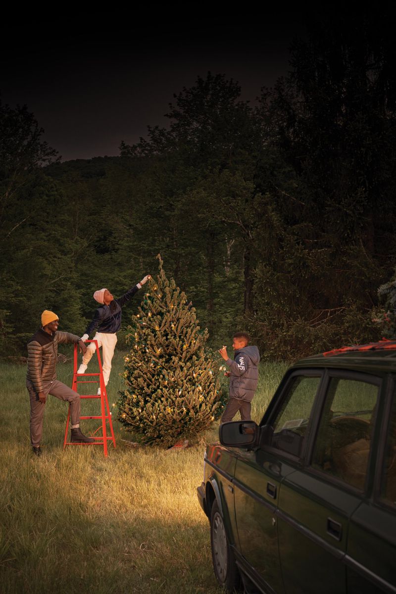 Three people decorating a Christmas tree outdoors at night near a car, illuminated by car headlight and string lights on the tree.
