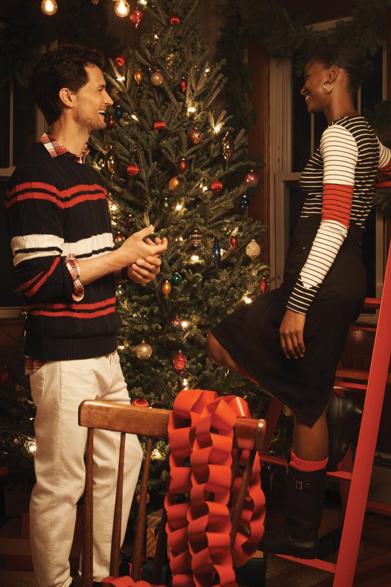 A couple decorates a Christmas tree in a warmly lit indoor scene. A man stands next to a woman on a ladder with holiday decorations.