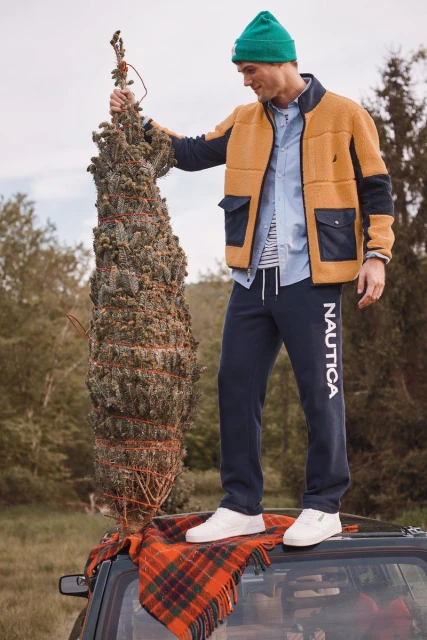 Adult male stands on a car roof in a green beanie, holding a Christmas tree with a plaid blanket and an outdoor background.