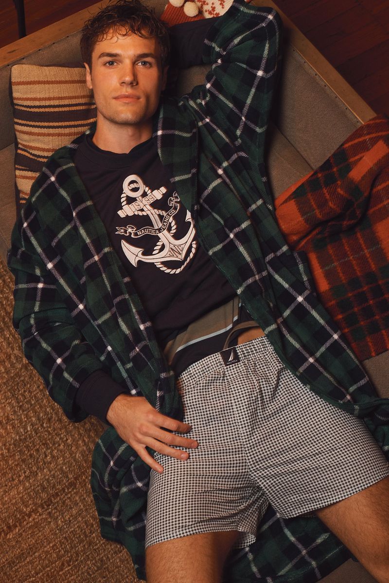 Man lying on a couch wearing underwear, a t-shirt, and a robe. He's indoors on a neutral couch with a patterned blanket.