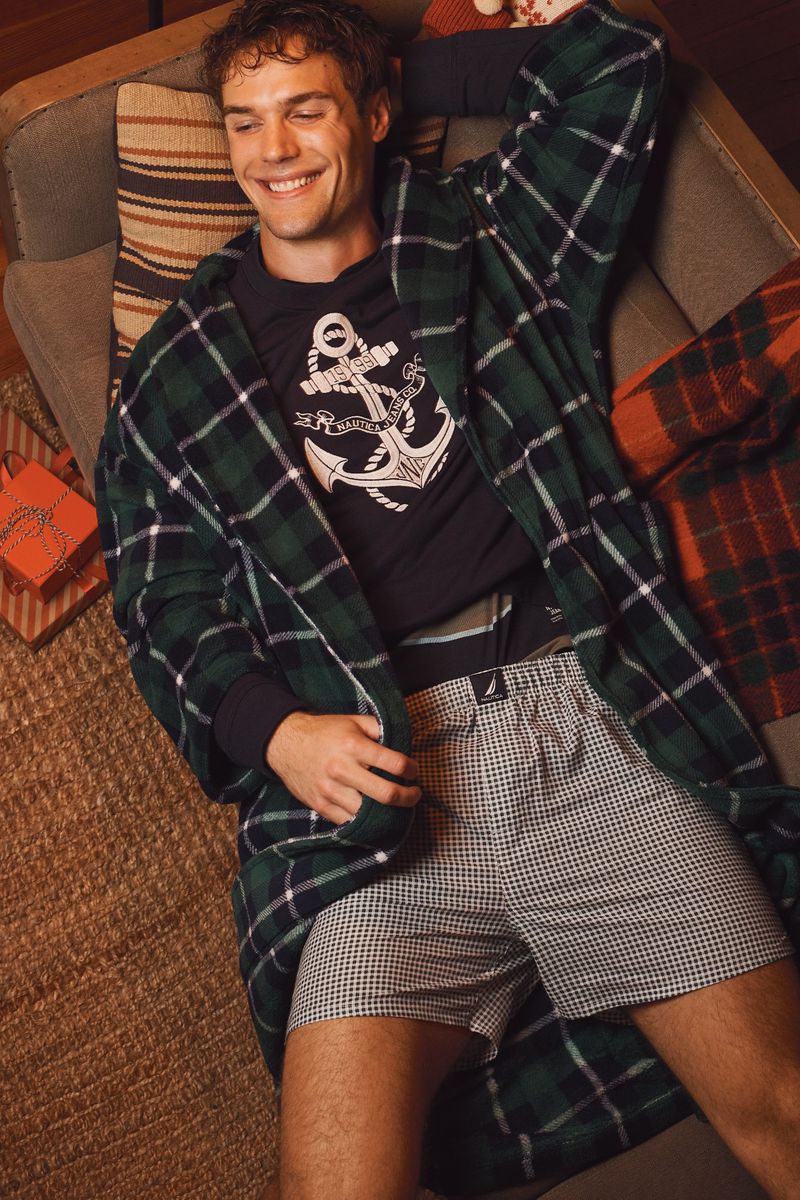 Smiling man lounges wearing plaid robe, black shirt with anchor, and patterned shorts, next to a striped pillow and wrapped gifts.
