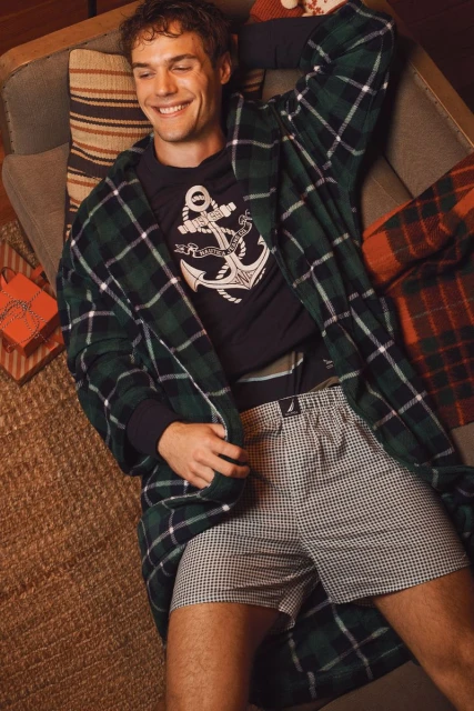 Smiling man lounges wearing plaid robe, black shirt with anchor, and patterned shorts, next to a striped pillow and wrapped gifts.