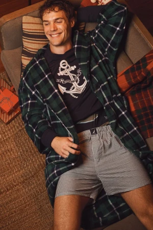 Smiling man lounges wearing plaid robe, black shirt with anchor, and patterned shorts, next to a striped pillow and wrapped gifts.