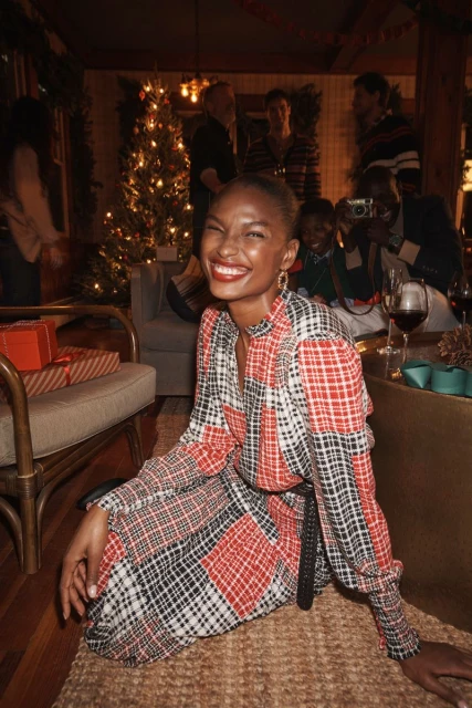 Smiling woman in a plaid dress sits on a rug in a festive living room with a Christmas tree and presents among a group of people.