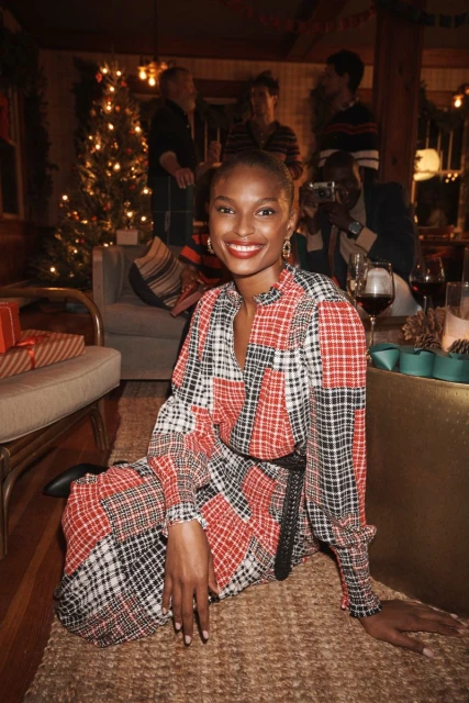 A woman with dark skin and red lipstick poses indoors near a Christmas tree, wearing a long-sleeved patchwork dress with a black belt and black boots.