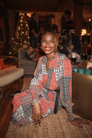 A woman with dark skin and red lipstick poses indoors near a Christmas tree, wearing a long-sleeved patchwork dress with a black belt and black boots.