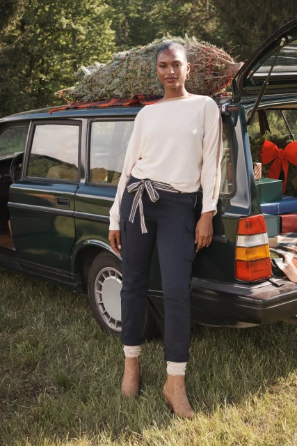 Woman in cargo pants, sweater, and booties stands with a Christmas tree in the back of a dark green vintage station wagon outdoors.