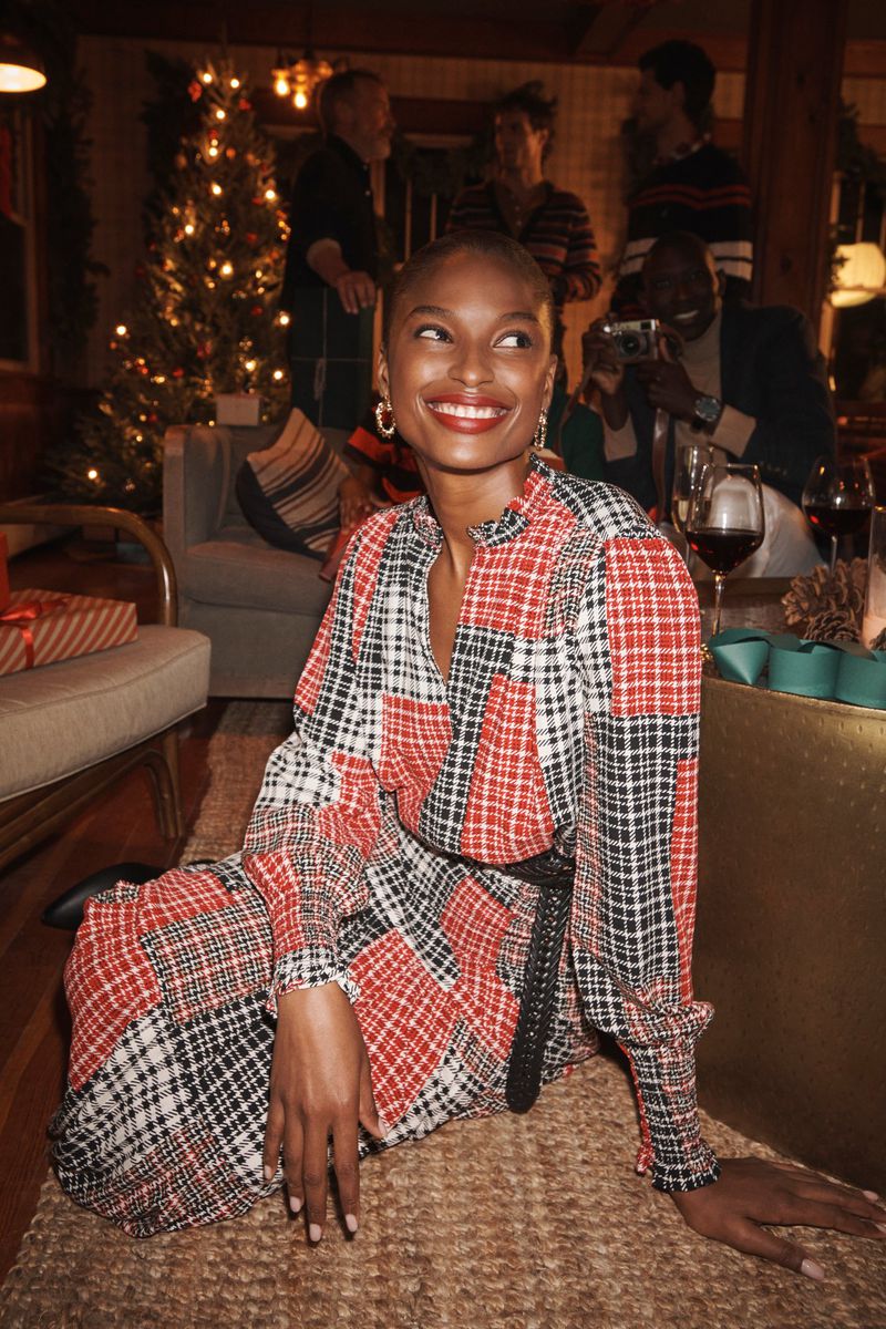 An African American woman in a plaid dress poses at an indoor Christmas party, surrounded by festive decorations and people celebrating.