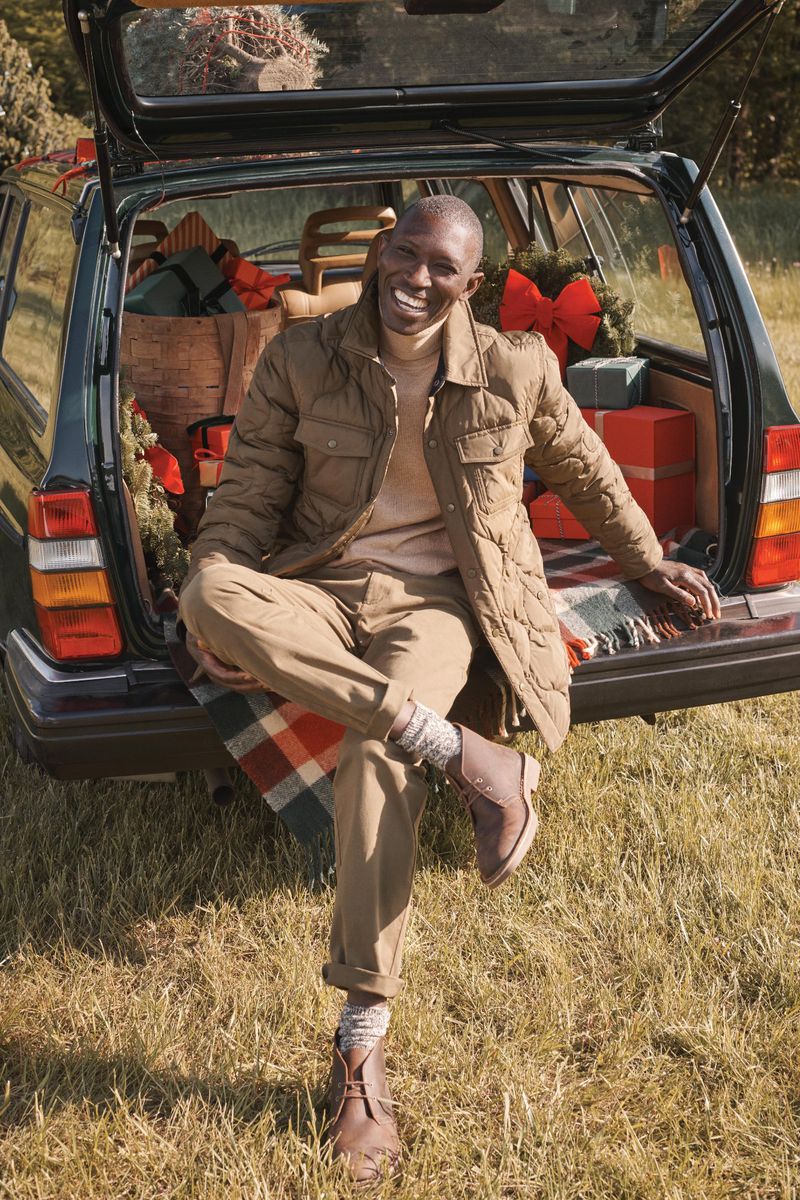 An African American man wearing a quilted jacket sits on the open trunk of a car filled with gifts on a grassy field in the daytime.