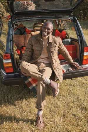 An African American man wearing a quilted jacket sits on the open trunk of a car filled with gifts on a grassy field in the daytime.