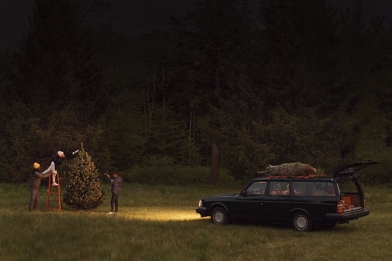 Three people decorate a Christmas tree near a vintage car in a forest at night, illuminated by a car headlight and Christmas tree lights.