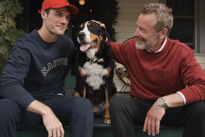 Two men with a Bernese Mountain Dog sit on a porch. One man wears a red cap and blue sweater; the other a red sweater and watch.