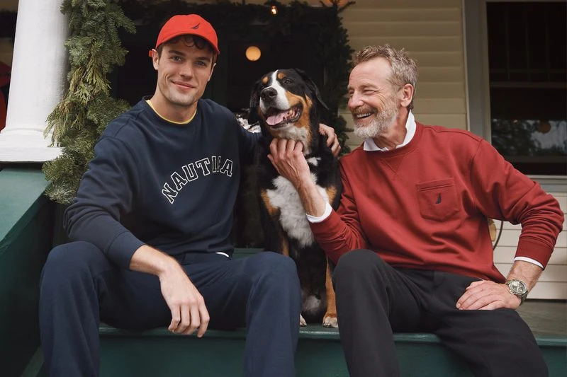 An older man and a young adult wearing sweaters pose with a dog on porch steps decorated with greenery and white lights. 
