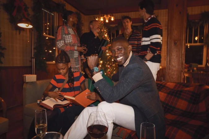 Group of people gather in a warmly decorated room, centered around a Christmas tree, with a smiling man in the foreground.