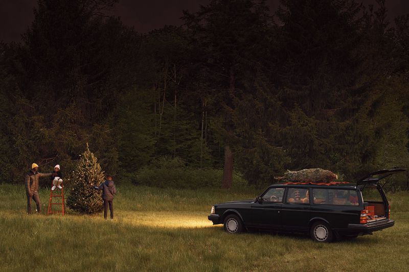 Family decorates Christmas tree outdoors in a field near a car with a tree on top, silhouetted against a dark forest and sky at night.