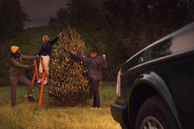 Family decorates a Christmas tree in a field with a vintage car in the background, using a ladder and stringing lights, at dusk.
