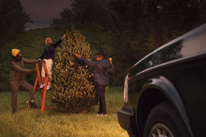 Family decorates a Christmas tree in a field with a vintage car in the background, using a ladder and stringing lights, at dusk.