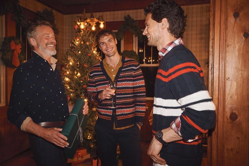 Three adult men smile in an indoor gathering near a Christmas tree, one holding a wrapped gift, creating a festive, convivial atmosphere.