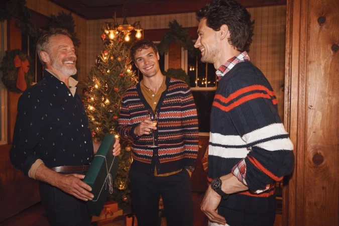 Three adult men smile in an indoor gathering near a Christmas tree, one holding a wrapped gift, creating a festive, convivial atmosphere.