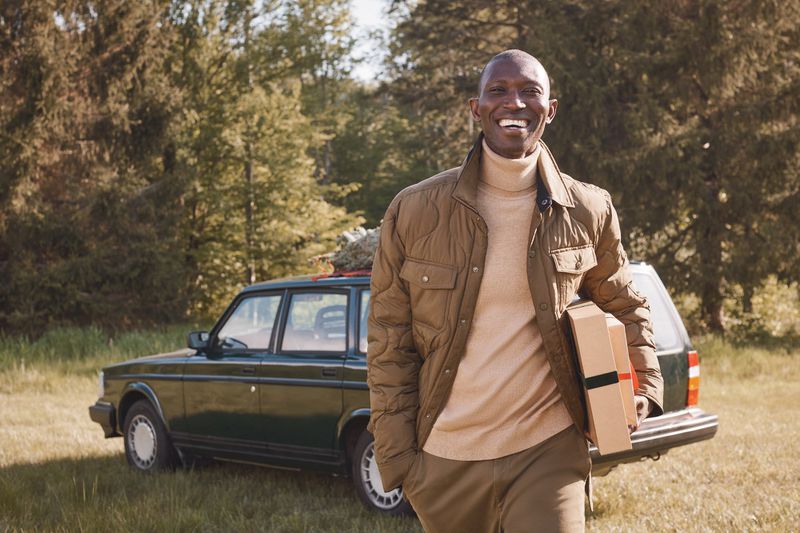 Smiling African American man carries a box, standing near a dark green Volvo car in a sunny field with trees in the background.