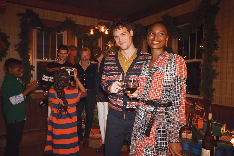 Group of adults and children in festive indoor setting. Diverse group poses with red wine and presents in a cozy, warmly lit room.