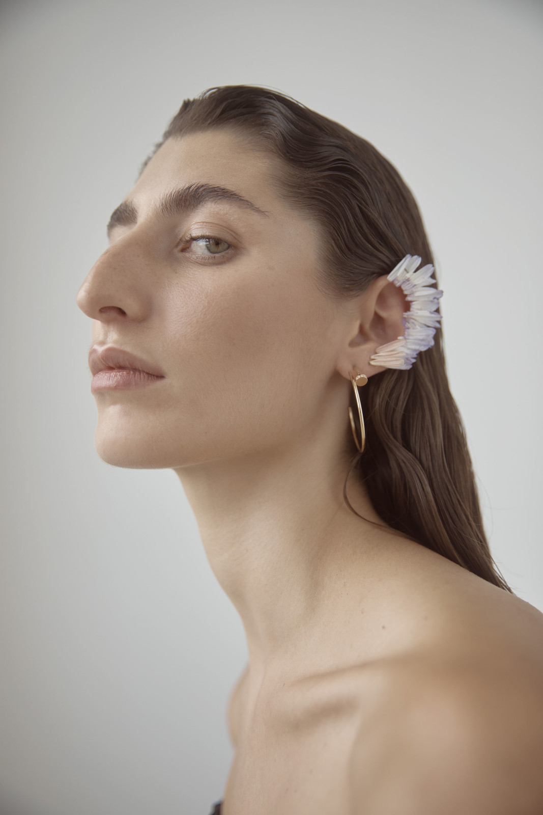Close-up portrait of a woman wearing an iridescent crystal ear cuff against a plain white background