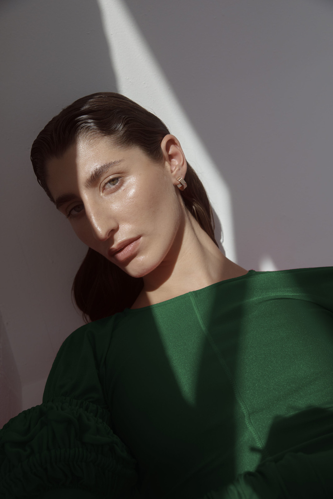 Portrait of an adult woman with dark hair wearing a green top, illuminated by a light with shadows, against a white backdrop.