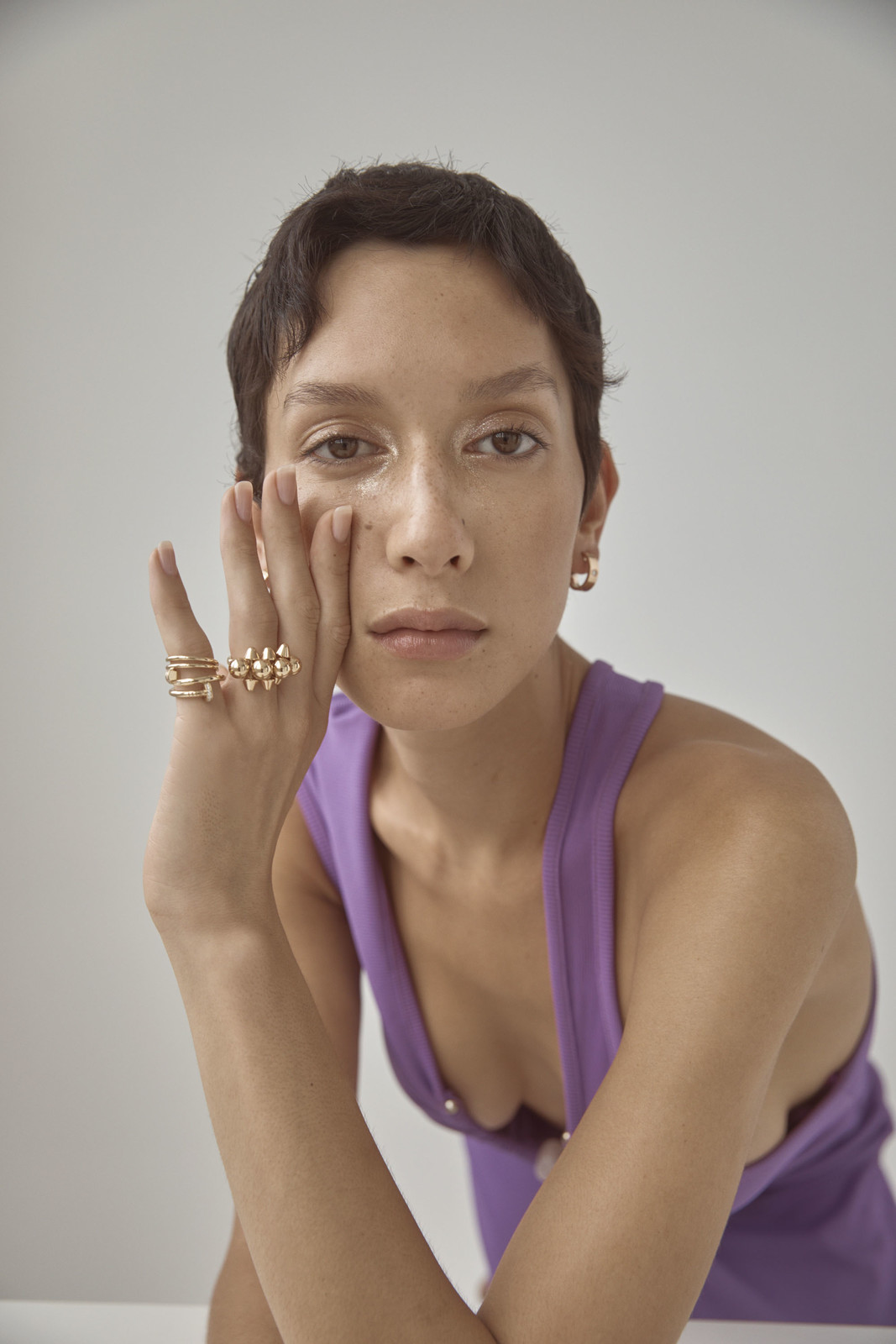 Close-up portrait of a woman with short brown hair wearing a purple tank top and gold jewelry, posing in front of a plain background.