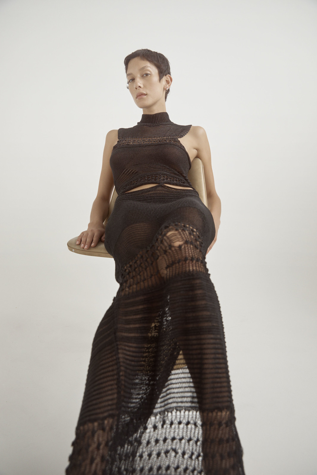 Model wearing a dark open-weave knit dress seated on a chair against a plain white background for Love Want Issue 24.