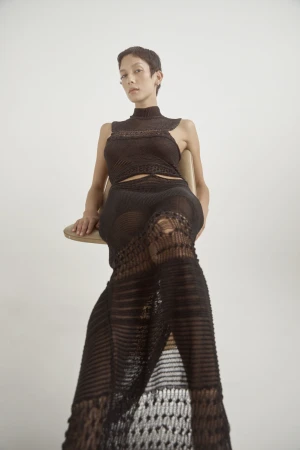 Model wearing a dark open-weave knit dress seated on a chair against a plain white background for Love Want Issue 24.